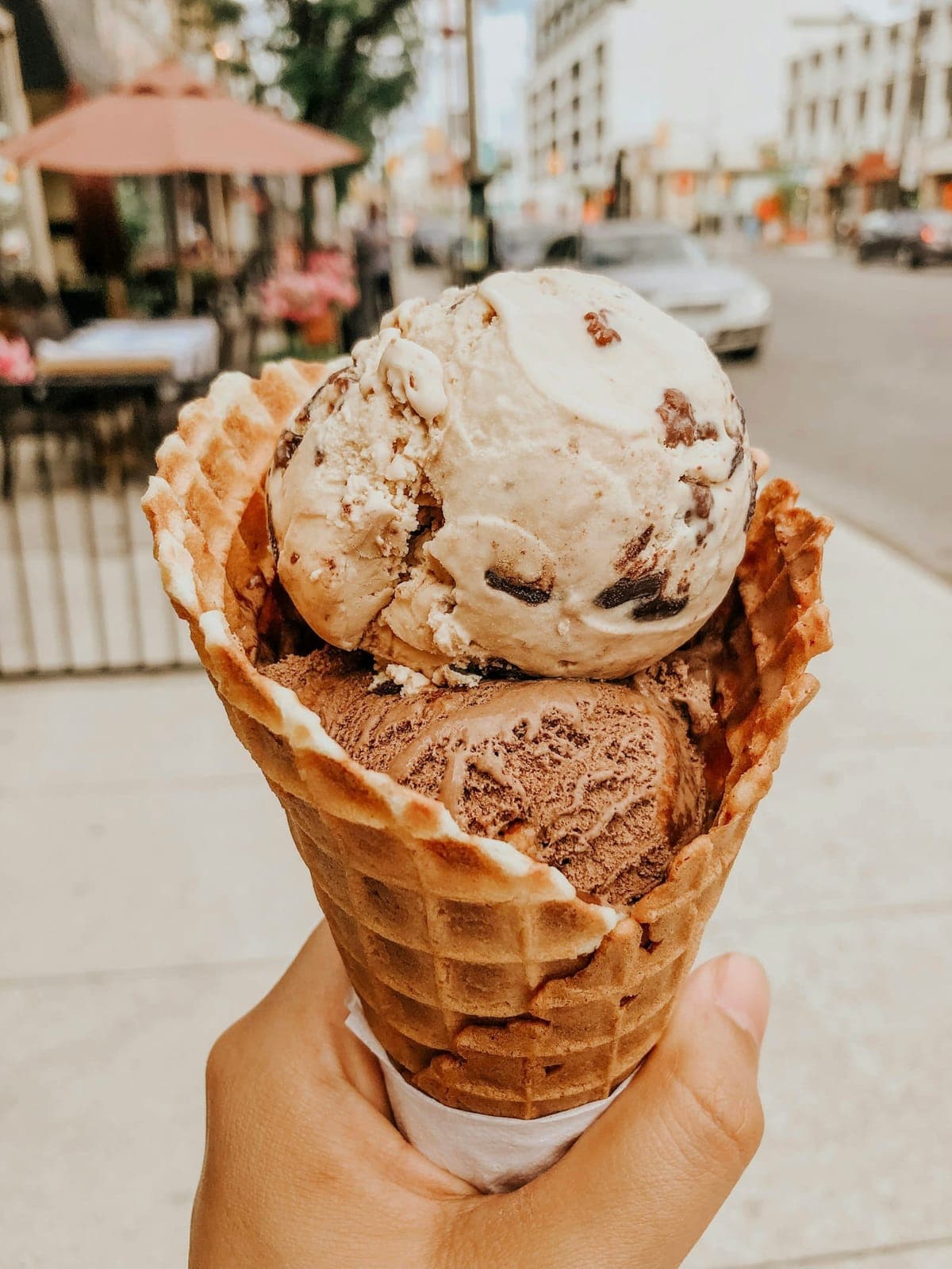 A double-scoop waffle cone held up on the sidewalk outside the stand